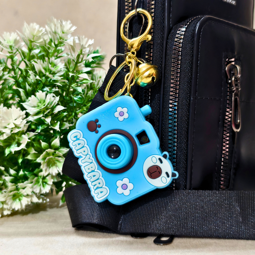 Blue capybara-shaped camera keychain attached to a black bag with green plants in the background