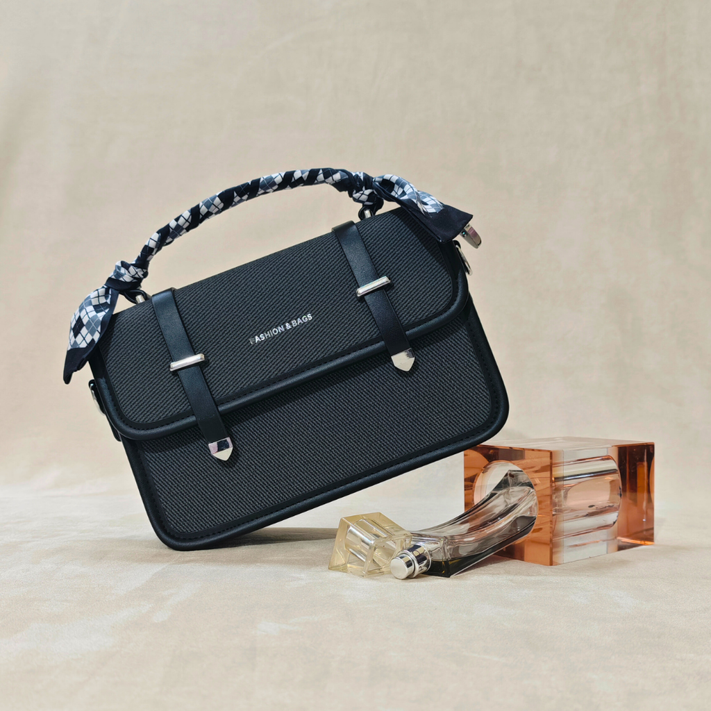 black sling bag in front of a beige background on top of a glass frame alongside a glass perfume bottle.