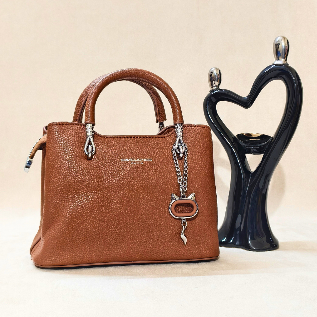 Brown handbag with a decorative charm next to a black heart-shaped candle holder on a beige background