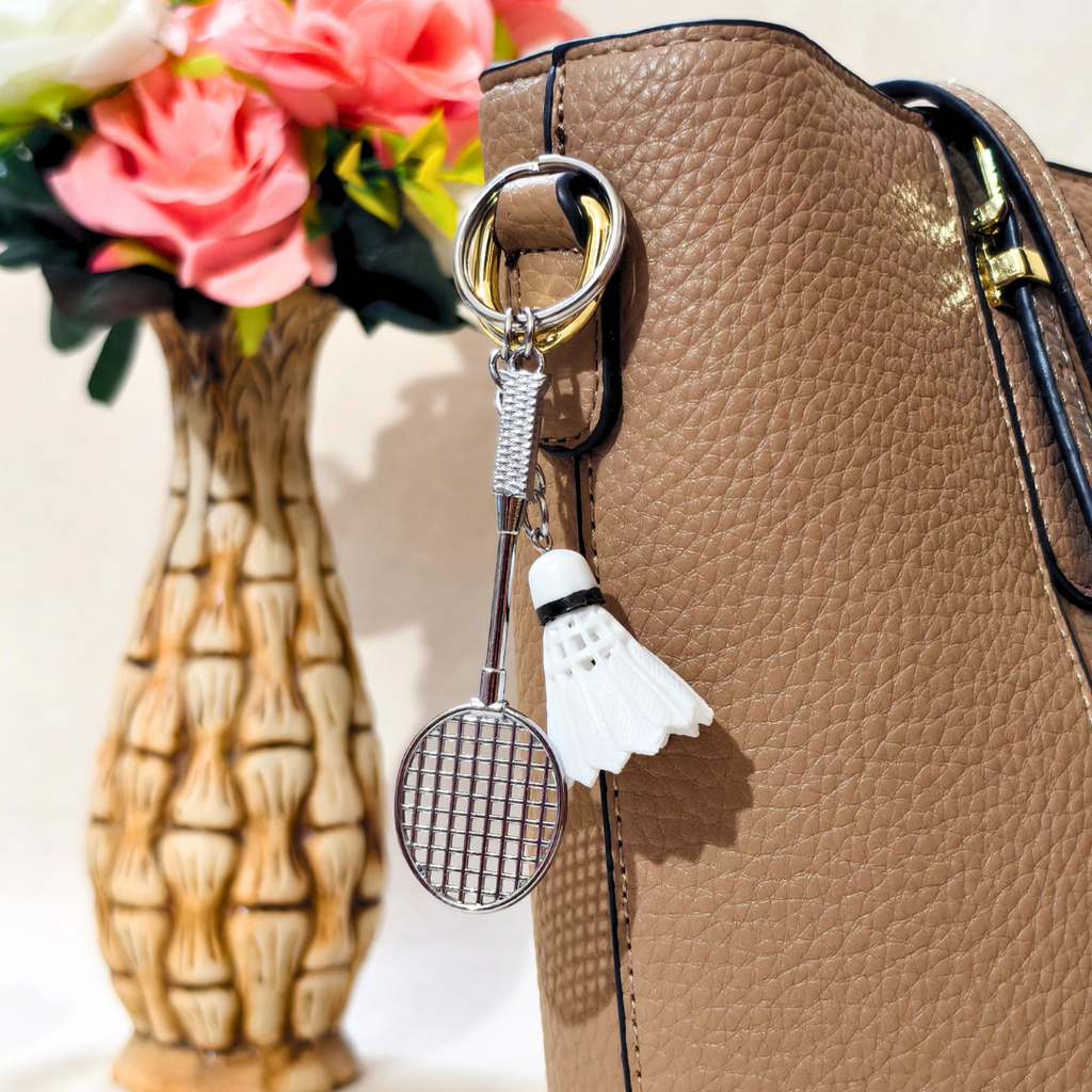 A close-up of a tan textured handbag with a silver metal ring and a white shuttlecock keychain charm hanging from it. A decorative beige vase with pink flowers is blurred in the background.