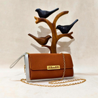 Brown clutch bag with gold chain and white accents on a beige background with wooden bird figurines.