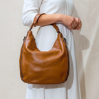 Brown leather handbag held by a person wearing a white dress against a plain background