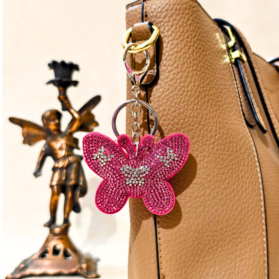 Pink butterfly keychain attached to a brown leather bag with a decorative figurine in the background.