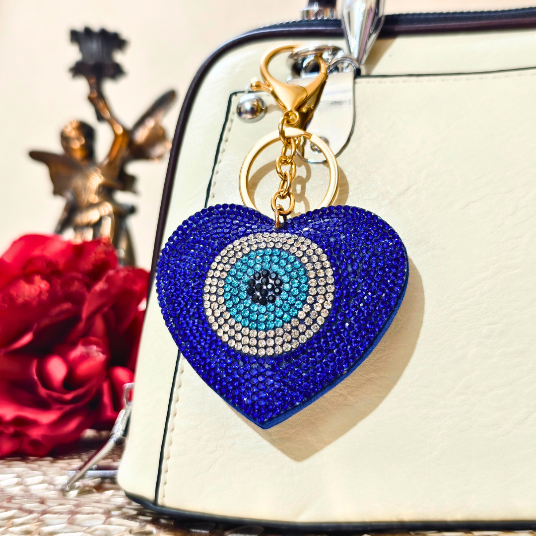 Blue heart-shaped keychain with eye design on a beige bag with red flowers and a gold figurine in the background.