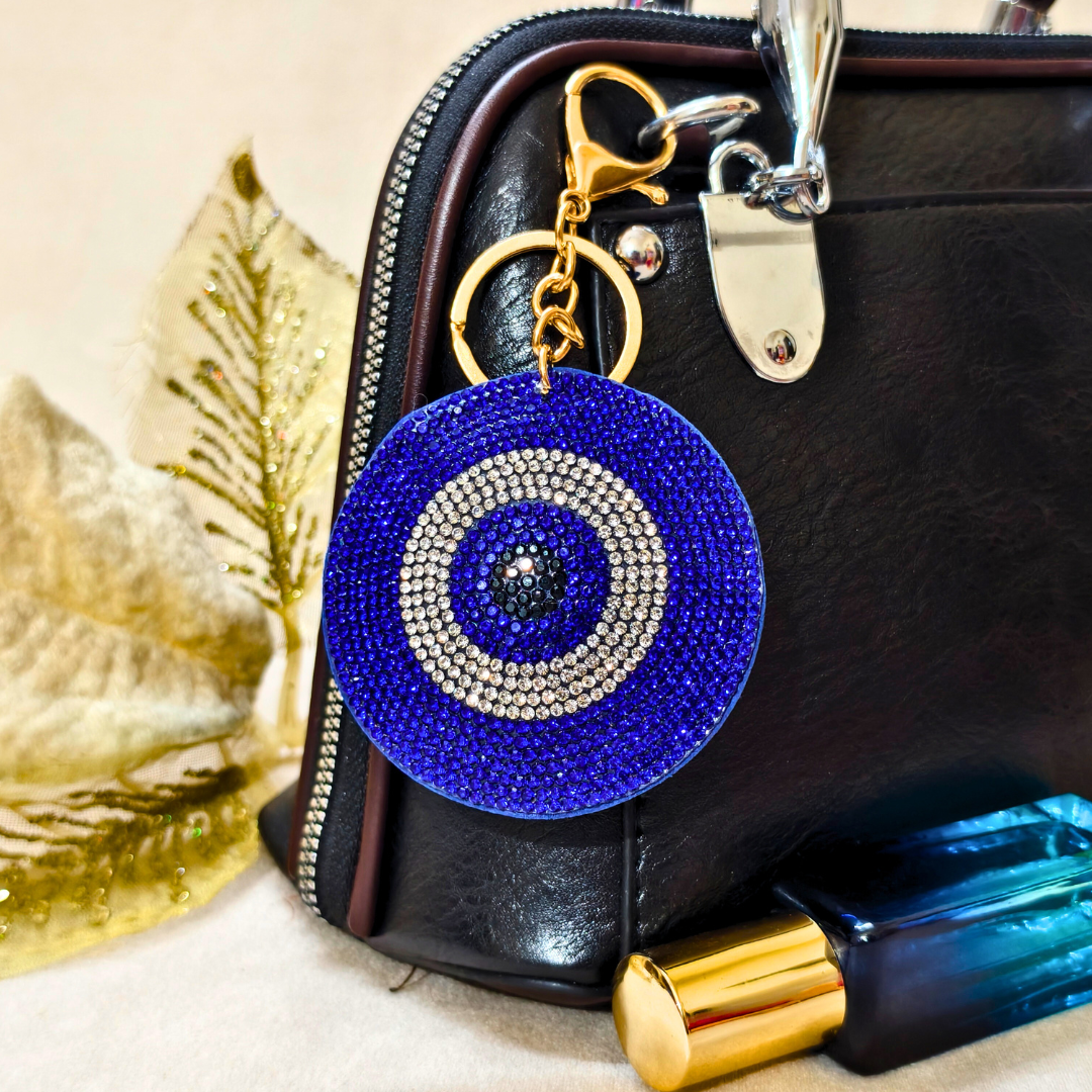 Blue evil eye keychain on a black handbag with a gold leaf and blue bottle in the background