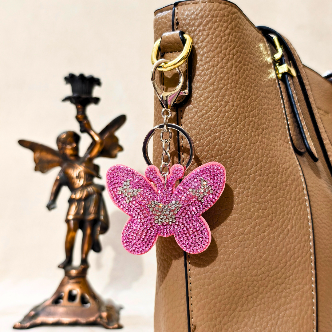 Pink butterfly keychain attached to a brown leather bag with a bronze angel figurine in the background.